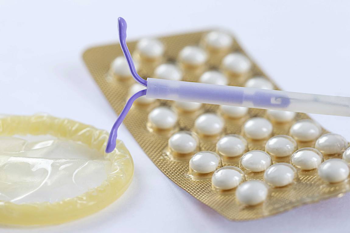 Nahaufnahme von drei Verhütungsmethoden: ein Kondom für Männer, eine Blisterpackung mit Antibabypillen und ein Intrauterinpessar (IUP) auf einer weißen Fläche.